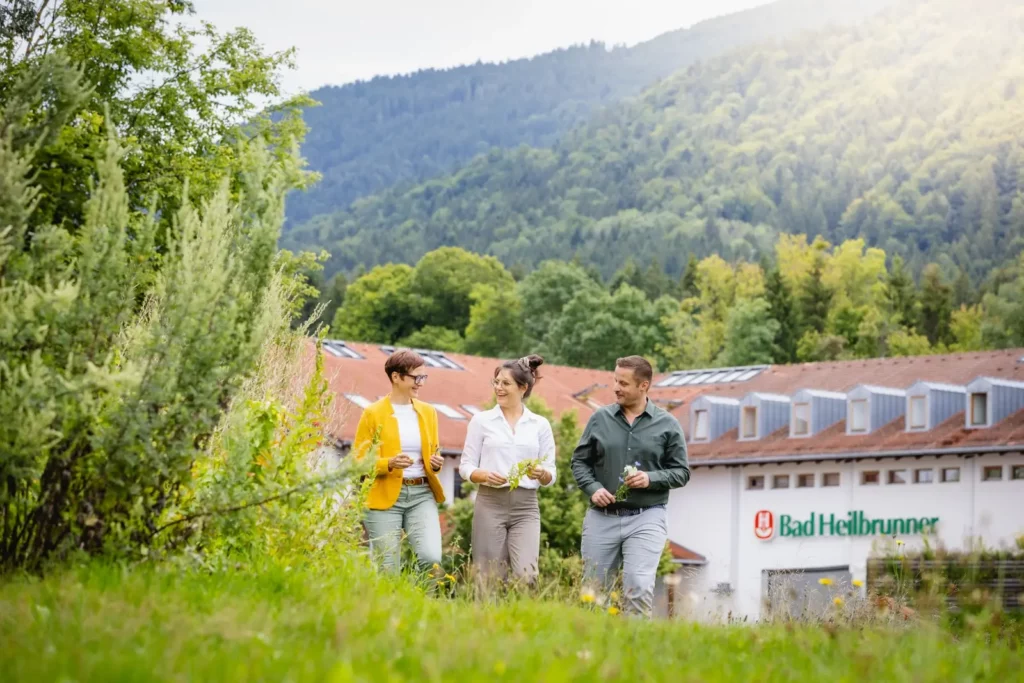 Drei Personen spazieren vor Bad Heilbrunner Gebäude in grüner Landschaft