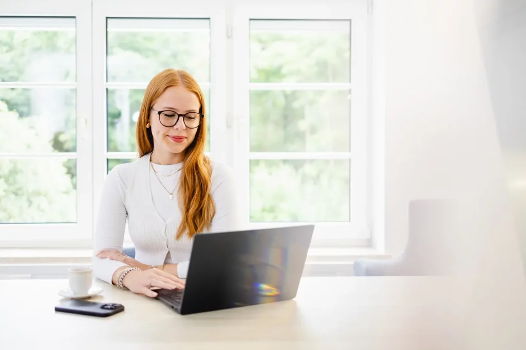 Rothaarige Frau arbeitet an einem Laptop vor einem Fenster