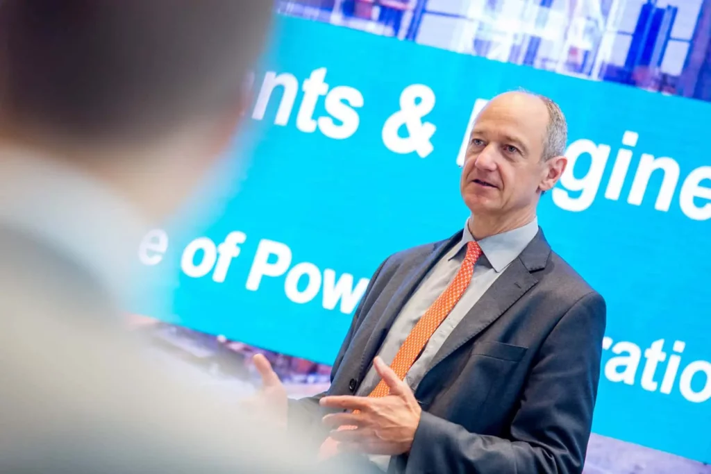 Mann im Anzug spricht vor Bildschirm mit Schriftzug