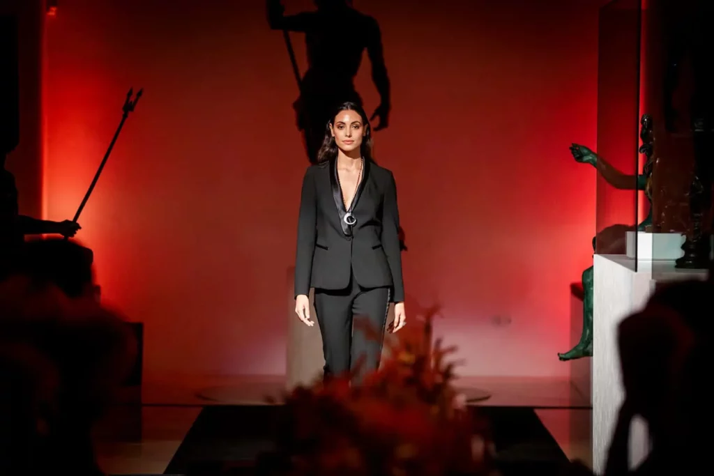 Frau in schwarzem Anzug läuft auf rotem Laufsteg mit dunkler Silhouette im Hintergrund