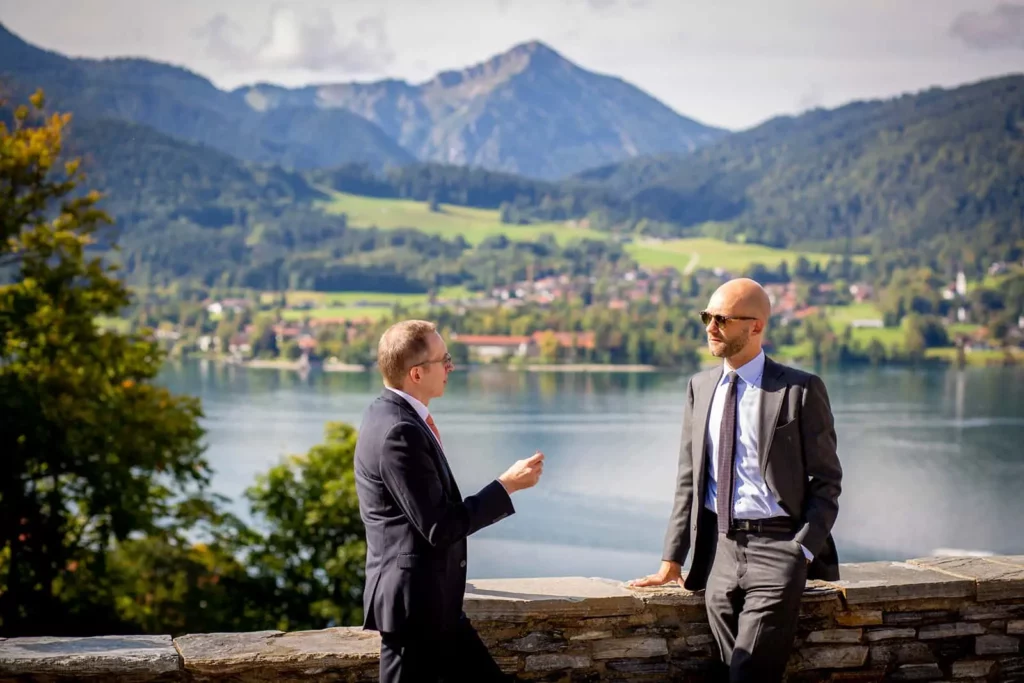 Zwei Männer in Anzügen sprechen vor einer Berg- und Seelandschaft