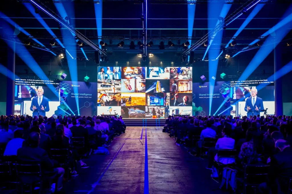 Konferenz mit Redner und großem Bildschirm, Publikum im Vordergrund