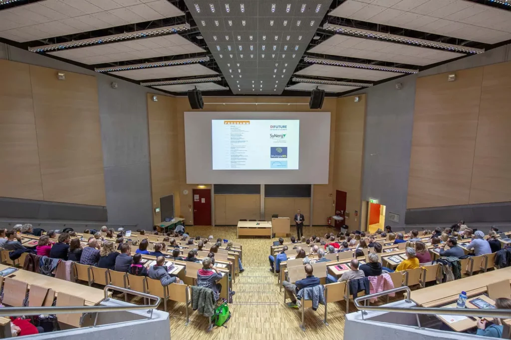 Gefüllter Hörsaal mit Studierenden und Dozent, Präsentation auf Leinwand