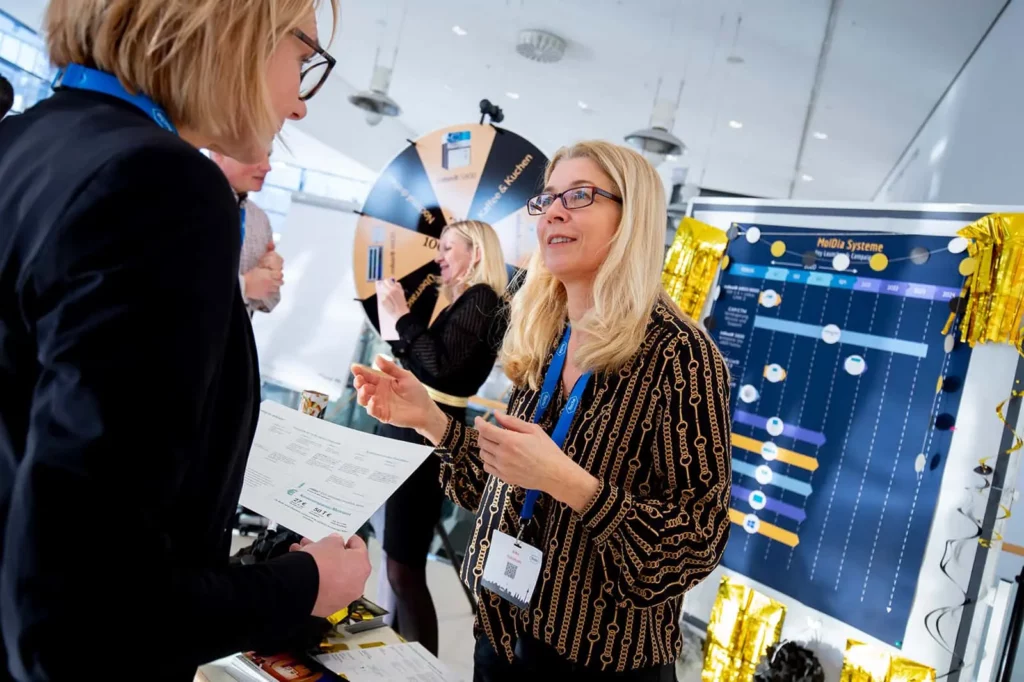 Geschäftsleute diskutieren vor Messestand mit Infotafel und Glücksrad
