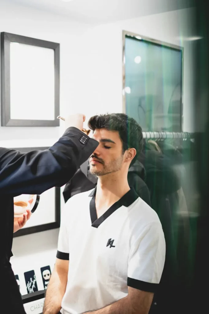 Mann im weißen Hemd wird vor einem Spiegel geschminkt