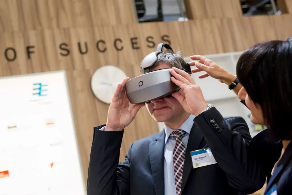 Mann testet Virtual-Reality-Headset auf Messe, Frau assistiert