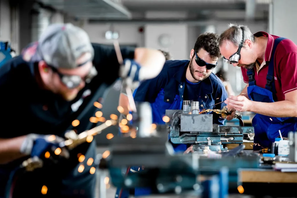 Drei Männer arbeiten in einer Metallwerkstatt mit Funken sprühenden Maschinen