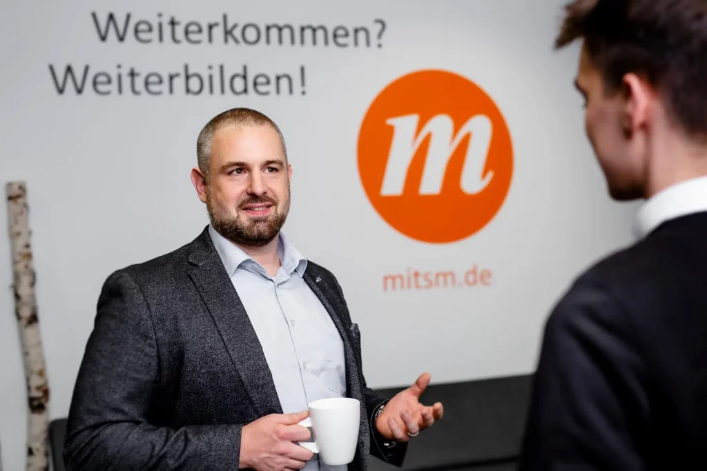 Geschäftsmann mit Tasse spricht mit Kollegen vor mitsm.de Logo