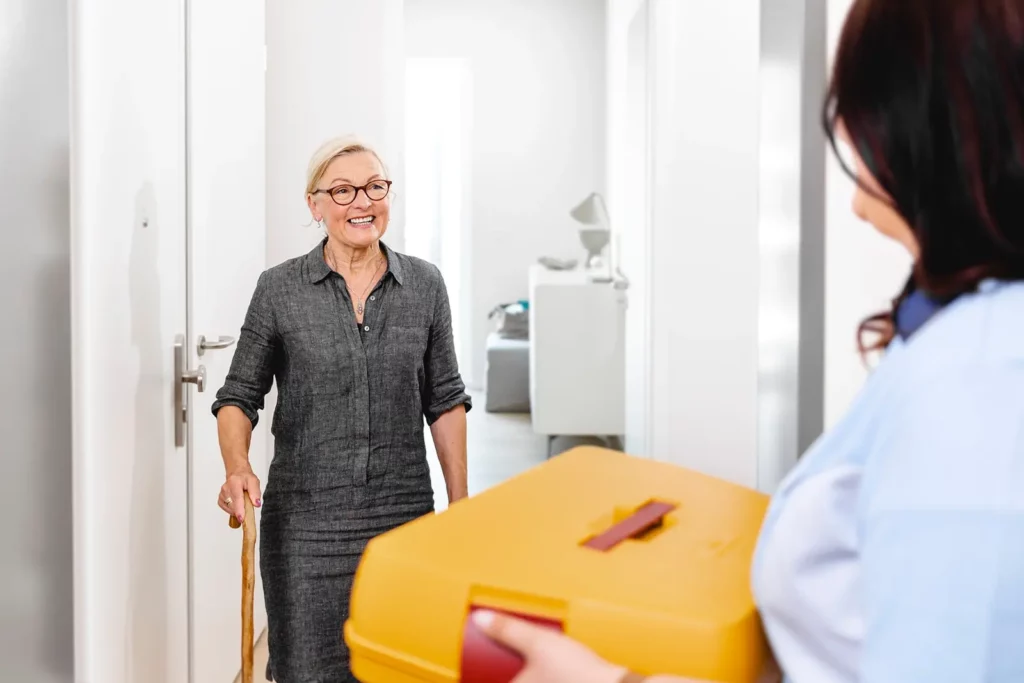 Seniorin öffnet glücklich die Tür für Pflegekraft mit gelber Box